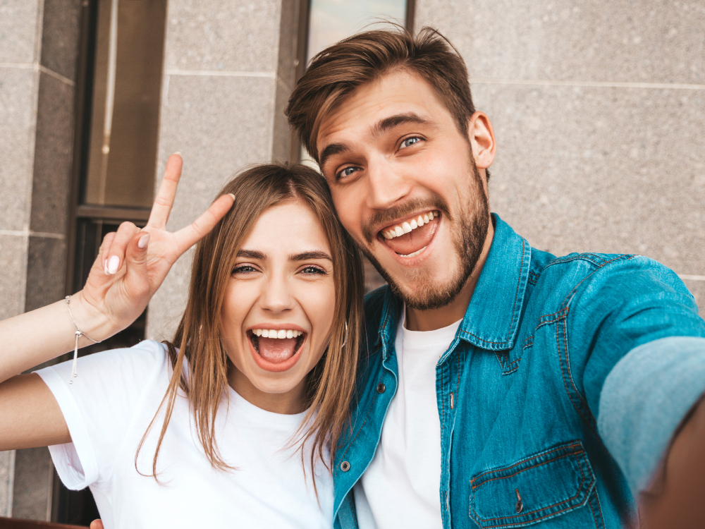 smiling-beautiful-girl-her-handsome-boyfriend-casual-summer-clothes-happy-family-taking-selfie-self-portrait-themselves-smartphone-camera-shows-peace-sign-winking-street